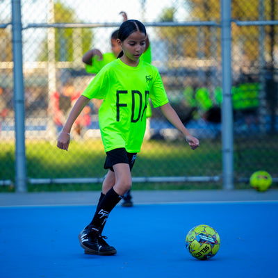 Child in a bright green Toque jersey playing soccer on a blue field with a fence in the background.