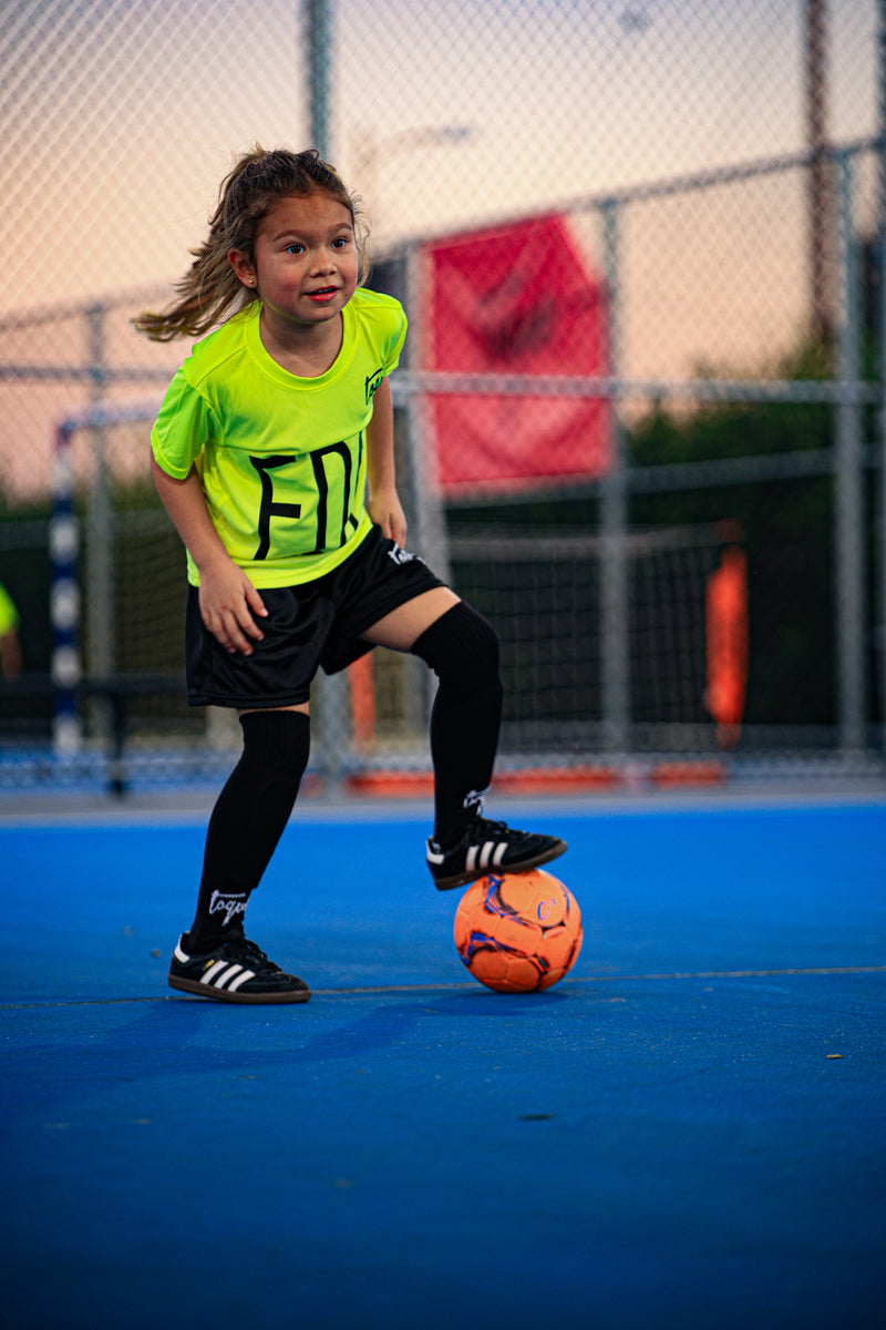 Young girl playing soccer on Toque field with a fence and sports equipment in the background.