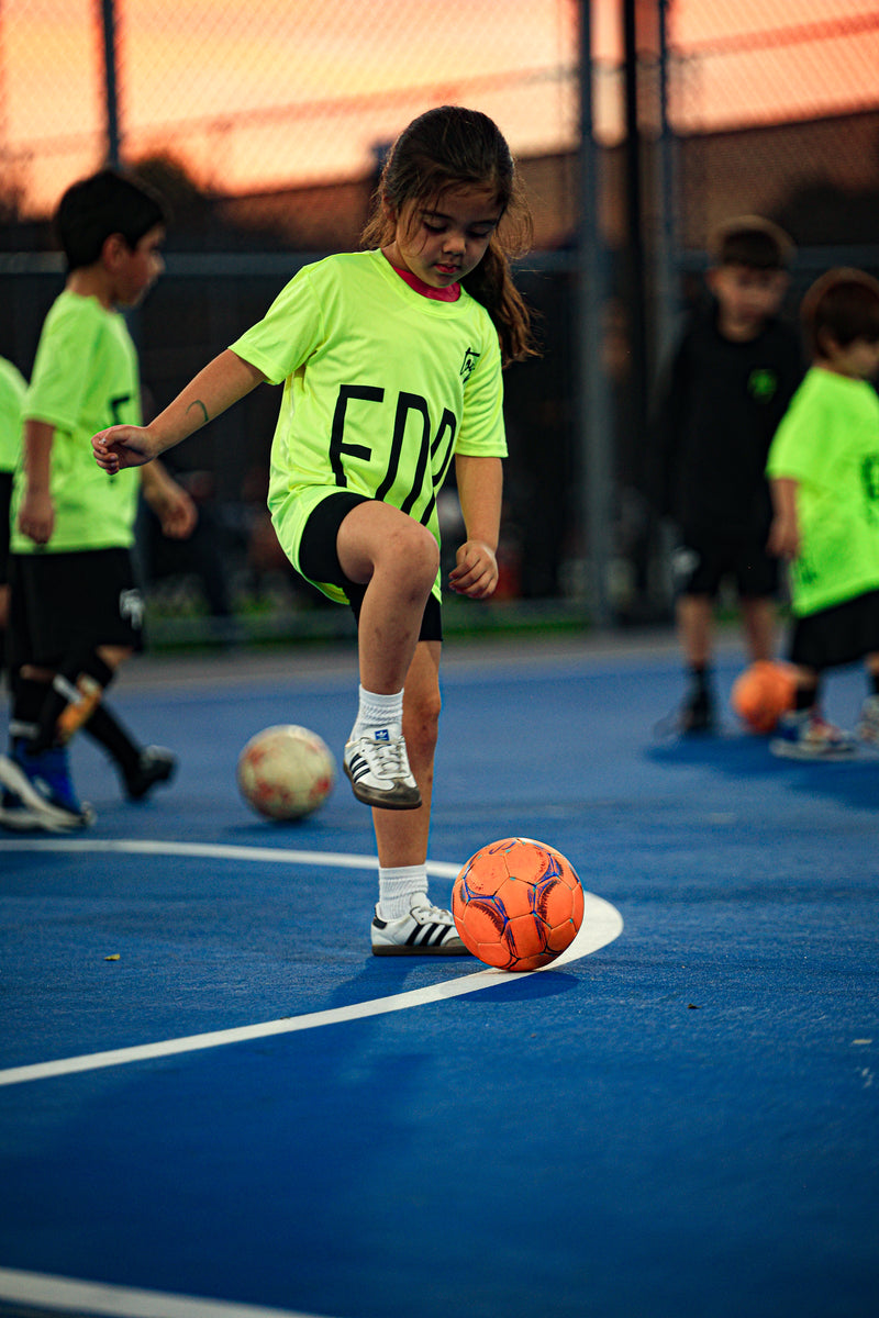 Child in a bright green Toque jersey kicking an orange soccer ball on a blue court.