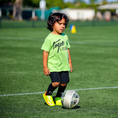Child in a green sports Toque jersey standing on a soccer field with a soccer ball.